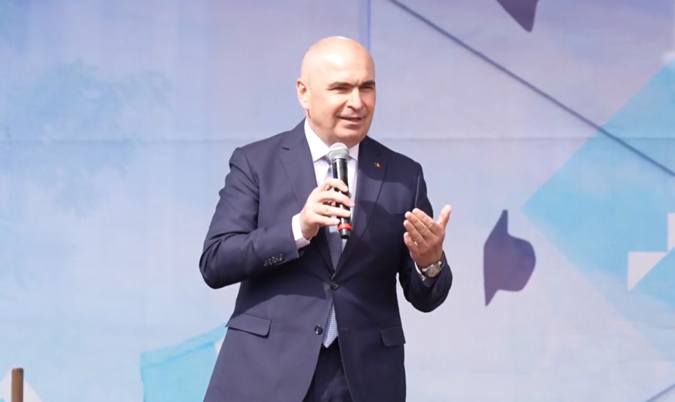 Man in a dark suit speaks into a handheld microphone on a stage with a blue abstract backdrop.