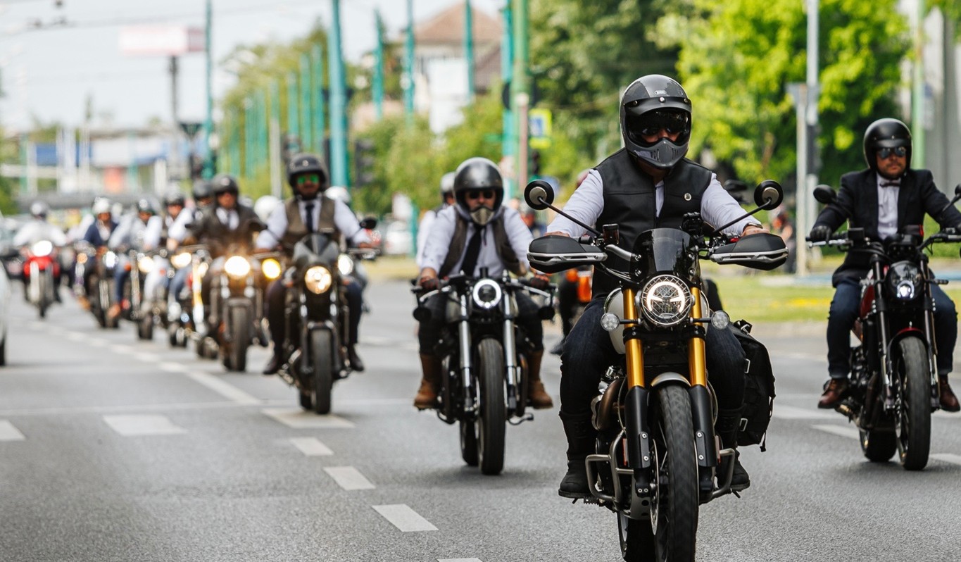 Group of motorcyclists riding down a city street, front rider wearing a black helmet and vest.