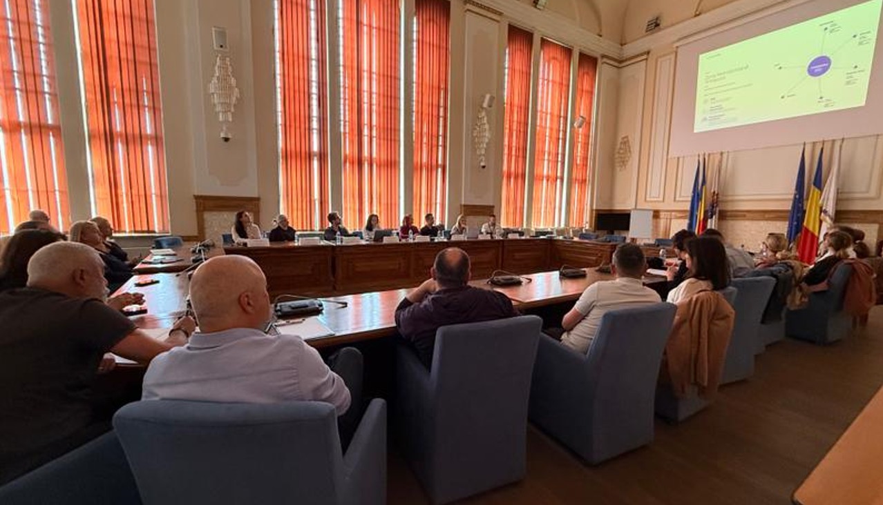 Formal meeting in a council chamber with people seated around a U-shaped wooden table and a large projector screen at the front.