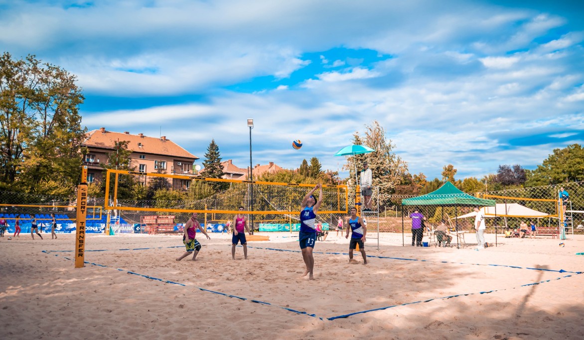 Beach volleyball match on a sunny sandy court with players jumping near the net and a ball in mid-air; spectators and tents in the background.