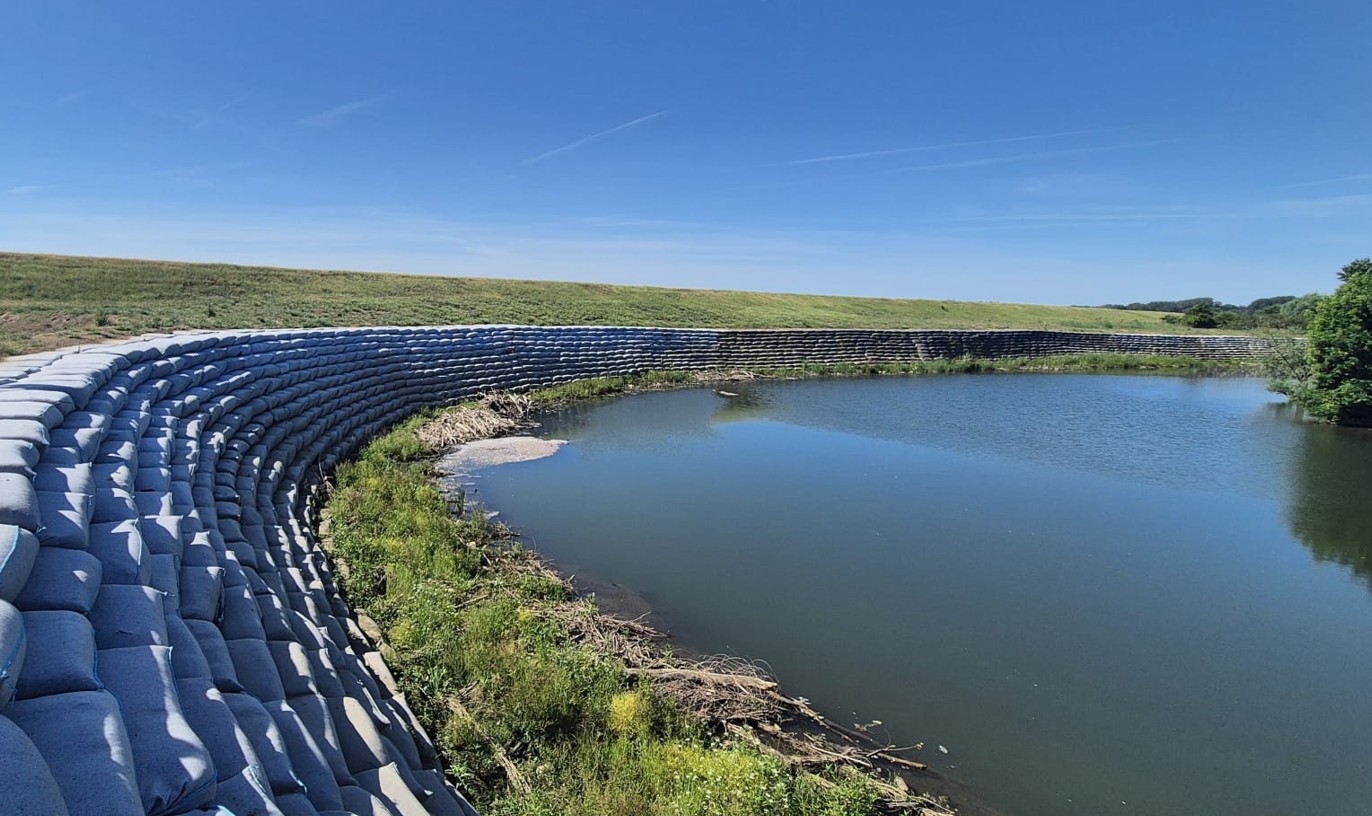 Curved wall of stacked concrete bags along a calm water body under a clear blue sky and grassy embankment beyond.