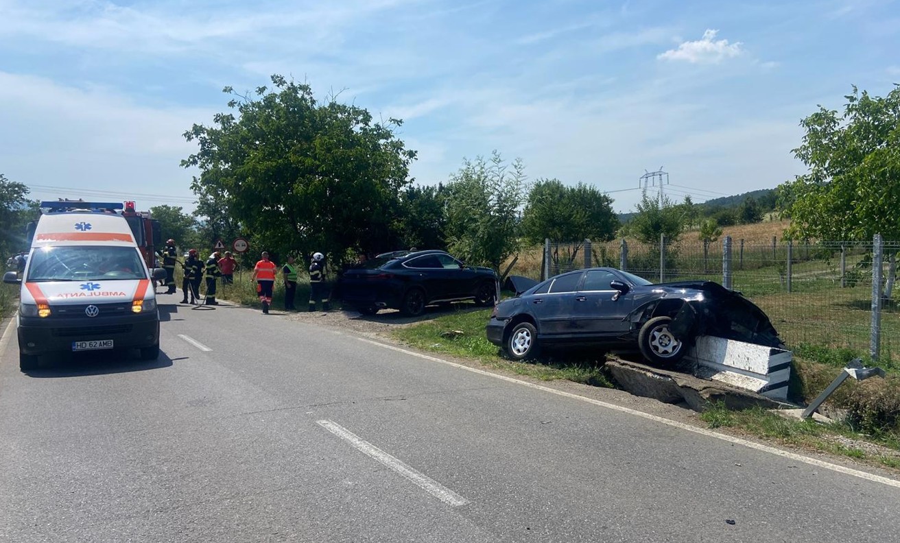 Ambulance and emergency responders on a sunny rural road at a two-car crash scene with one car off the road beside a fence.