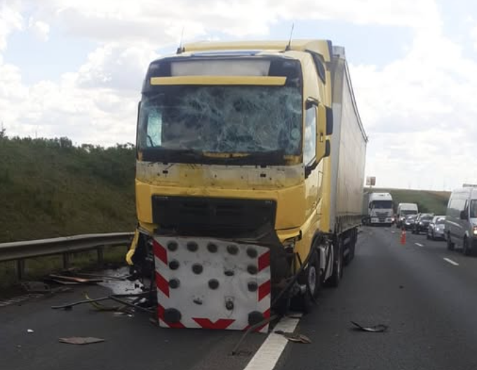 Yellow semi-truck with severe front-end damage on a highway, windshield cracked and debris scattered as traffic backs up nearby.