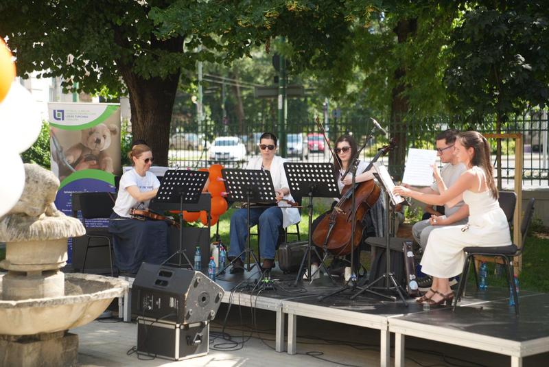 A small outdoor string ensemble performing on a portable stage in a park, with sheet music stands and a banner in the background.