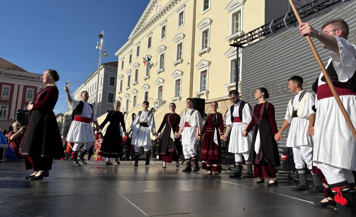 Group of people in traditional folk costumes performing a dance on an outdoor stage in front of a yellow historic building.