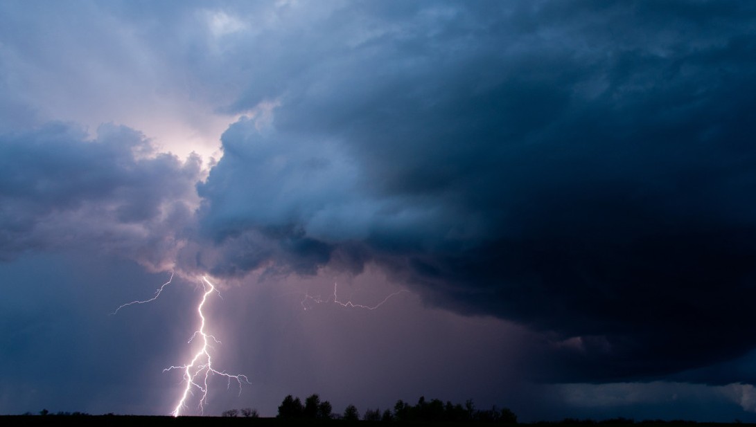 Lightning bolt streaks down through dark storm clouds over a flat horizon at dusk.