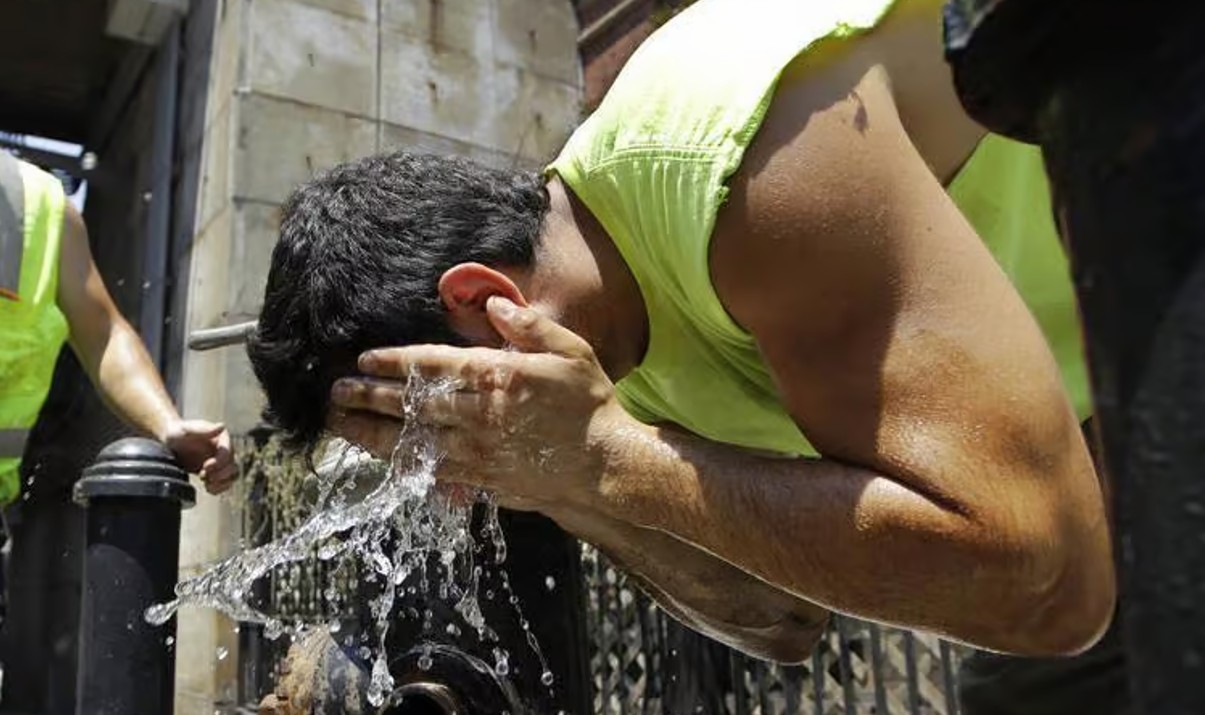 Man in a neon sleeveless shirt splashes water on his head from his hands at an outdoor water fountain, with a coworker in a safety vest nearby.