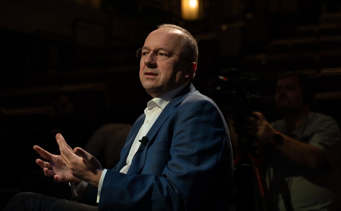 Man in a blue suit being interviewed, gesturing with his hands while speaking, in a dim studio with a camera operator in the background.