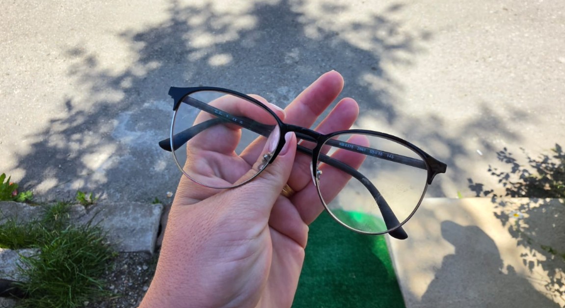 Hand holding black-framed eyeglasses with round lenses outdoors on a paved surface.