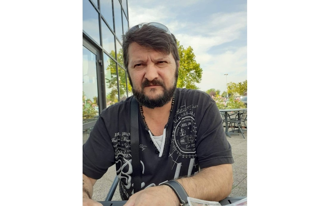 Bearded man wearing a black graphic shirt sits at an outdoor cafe, sunglasses on his head.