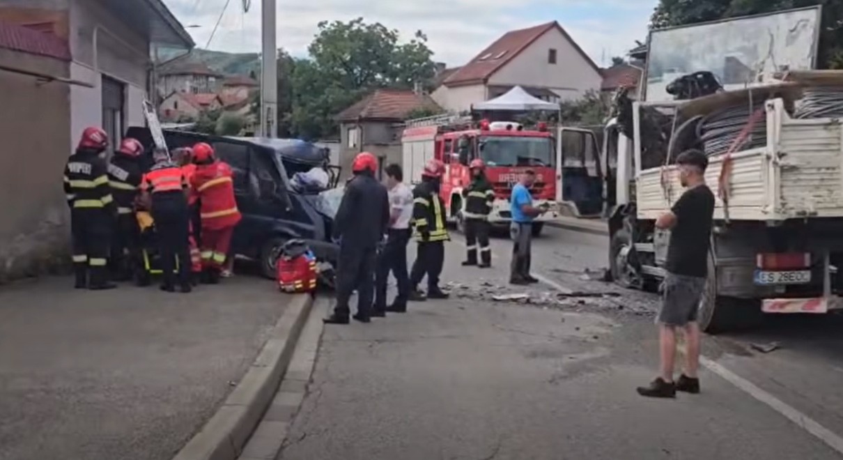 Firefighters and responders work at a car crash on a street with a tow truck and debris nearby, a fire truck in the background.