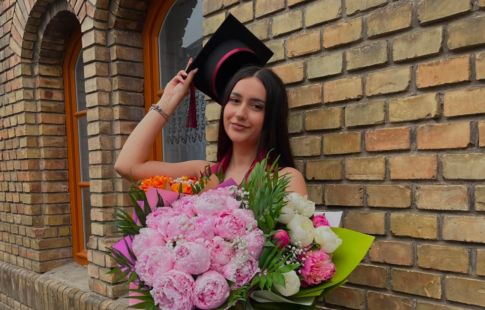 Graduate woman wearing a cap and gown holding a large bouquet of pink and white flowers outside a brick building.