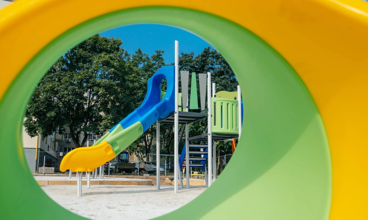 Colorful playground structure with a blue slide and green railings seen through a yellow circular tunnel outdoors, with trees and buildings in the background.
