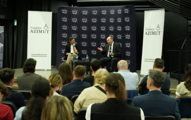 Two men on a stage in a panel discussion, audience seated in front, banners display Fundatia AZIMUT.