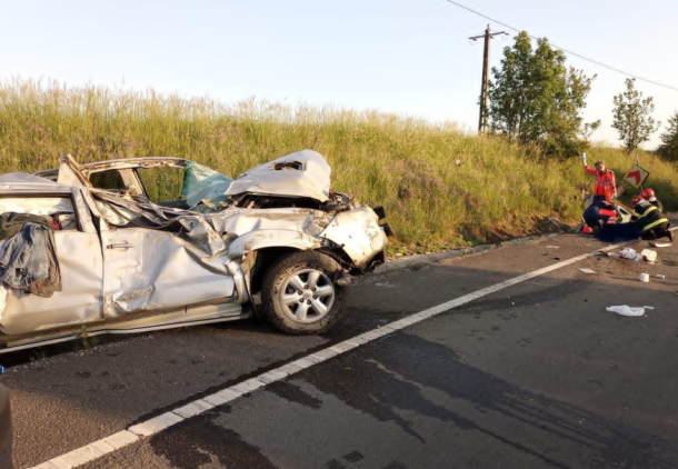 Wrecked white sedan after a severe crash on a road with emergency responders tending to a person nearby on the grass verge to the right of the scene.
