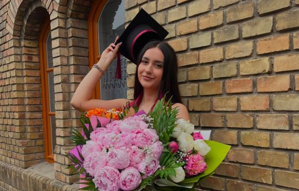 Graduate woman wearing a cap and gown holding a large bouquet of pink and white flowers outside a brick building.