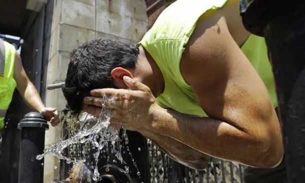 Man in a neon sleeveless shirt splashes water on his head from his hands at an outdoor water fountain, with a coworker in a safety vest nearby.