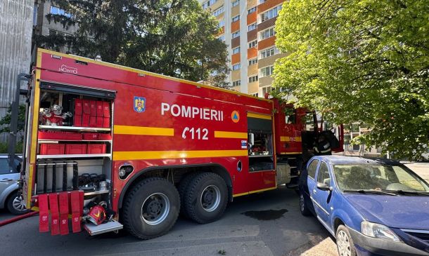 Red fire truck with yellow stripes labeled POMPIERII 112 parked on a city street beside a blue car with a residential building in the background and trees nearby.