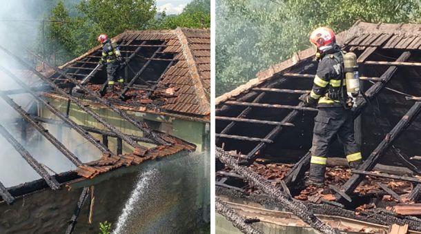 Firefighter in helmet and turnout gear on a heavily damaged tiled roof, directing a water spray as smoke rises from the burned house.