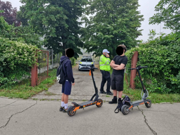 Two kids with orange-and-black scooters on a residential street as a police officer speaks nearby.