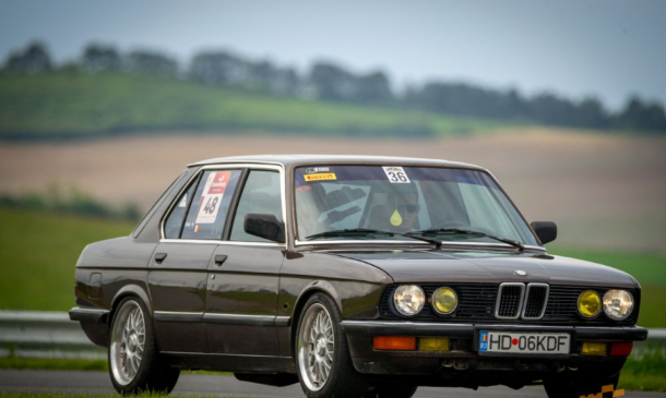 Brown 1980s BMW race car with numbered stickers on a track, front three-quarter view.