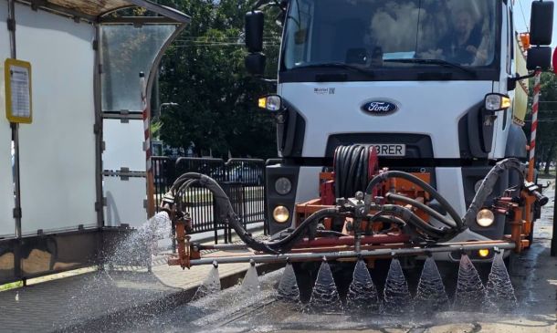 Front view of a white Ford street-cleaning truck spraying water through a bar with multiple nozzles onto the street surface.