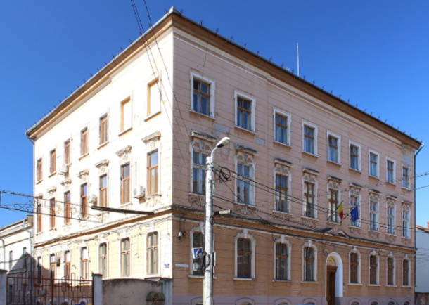 Three-story beige-orange building with decorative window frames and an arched entrance, flags by the facade, under a clear blue sky.