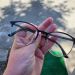 Hand holding black-framed eyeglasses with round lenses outdoors on a paved surface.