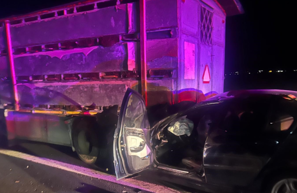 Nighttime crash scene: a black car has collided with the side of a large trailer truck, the driver's door is open and interior is visible with purple emergency lights.