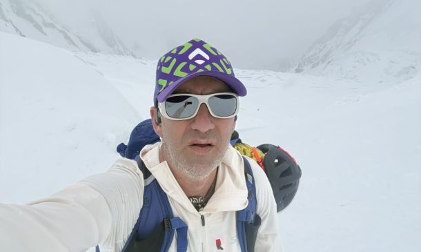 Man in a white jacket takes a selfie in a snowy, mountainous landscape wearing a purple-green cap and white sunglasses with a backpack behind him.