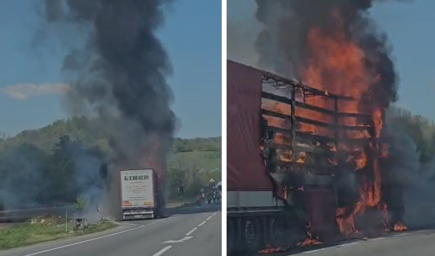 A white box truck on a two-lane road with a tall plume of thick black smoke rising from its rear, in a rural area with hills in the background.