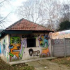 Small, graffiti-covered one-story building with a tiled, sloped roof and colorful cartoon faces on its walls, beside a path and fence.