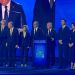 Group of formally dressed people on a stage at a conference, with a podium in the center.