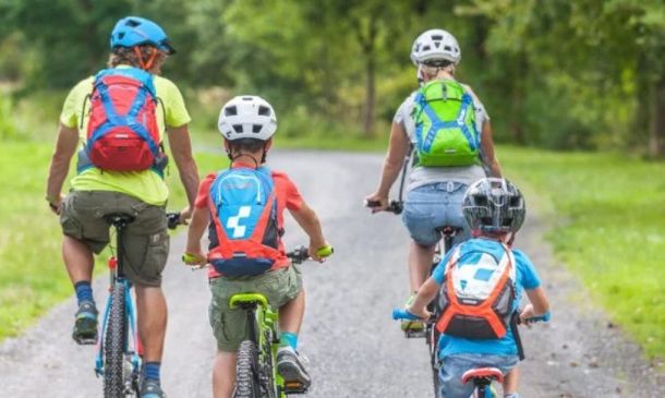 Four people (two adults and two children) ride bicycles on a rural path, wearing helmets and colorful backpacks.