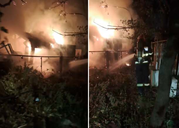 Nighttime yard fire with flames and smoke; a metal fence and debris, firefighters spraying water toward the blaze.