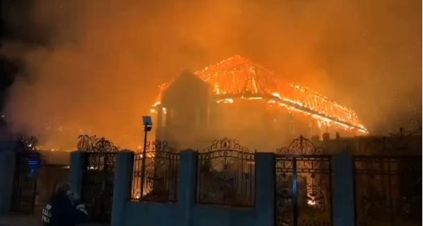House engulfed in flames at night, bright orange fire consuming the roof behind a decorative gate.