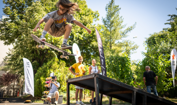 Skater launches off a ramp, mid-air, performing a kickflip while others watch in a sunny park.
