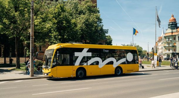 Yellow city bus with a large white logo on its side parked along a street with trees and buildings in the background.