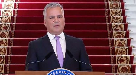 Man in a dark suit and lavender tie delivering a speech at a podium, with red-carpet stairs behind him.