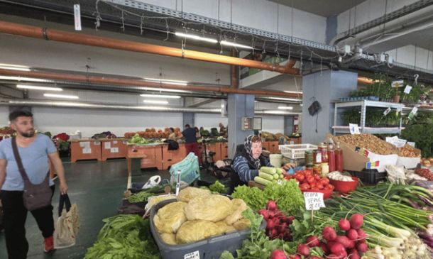 Indoor market stall with fresh vegetables displayed in crates; a vendor wearing headphones sits behind the produce while shoppers browse in the background.