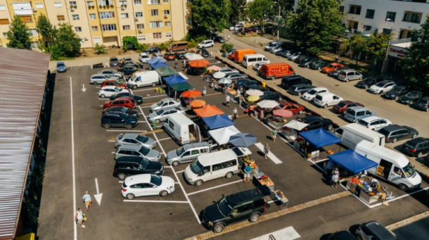 Overhead view of a bustling outdoor market in a parking lot, with colorful tents, vendors, and shoppers among parked cars and apartment buildings nearby.