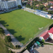 Aerial view of a green rectangular soccer field with goal nets, surrounded by bleachers and buildings. Nearby houses line the hillside beyond the stadium.