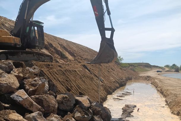 Excavator on a rocky slope digging a long trench along a waterline, with muddy banks and a cloudy sky.