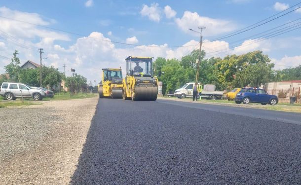 Two yellow road rollers compacting fresh asphalt on a rural street, with parked cars and workers in the background.