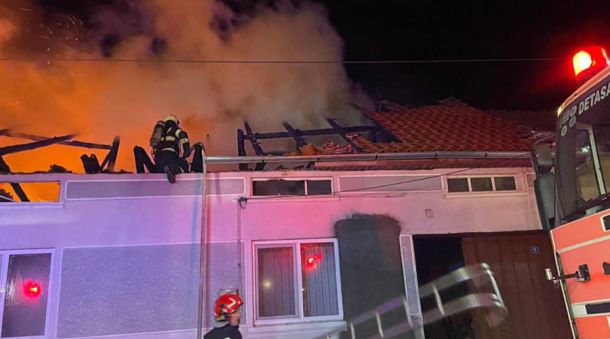 Firefighters on a burning building roof at night as flames and thick smoke rise, with a fire engine in view at the right. (Informative)
