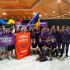 Group of students in purple team shirts posing with banners and a Ukrainian flag at a robotics competition, trophies on the floor, indoor arena backdrop