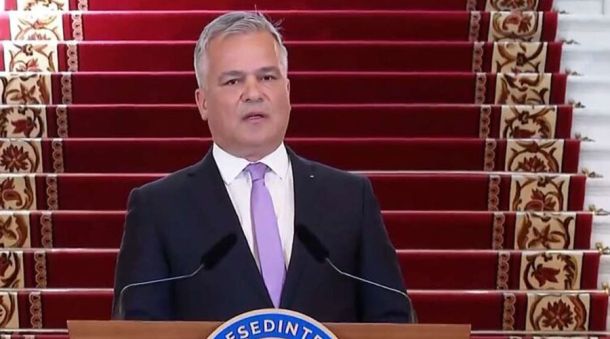 Man in a dark suit and lavender tie delivering a speech at a podium, with red-carpet stairs behind him.