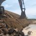 Excavator on a rocky slope digging a long trench along a waterline, with muddy banks and a cloudy sky.