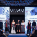 Runway fashion show: models in diverse outfits stand on stage under blue lighting; audience watches, with large screens showing text and images behind them.