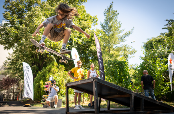 Skater launches off a ramp, mid-air, performing a kickflip while others watch in a sunny park.